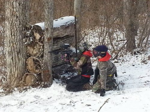 2 boys in snow shooting paintball gun from behind logs